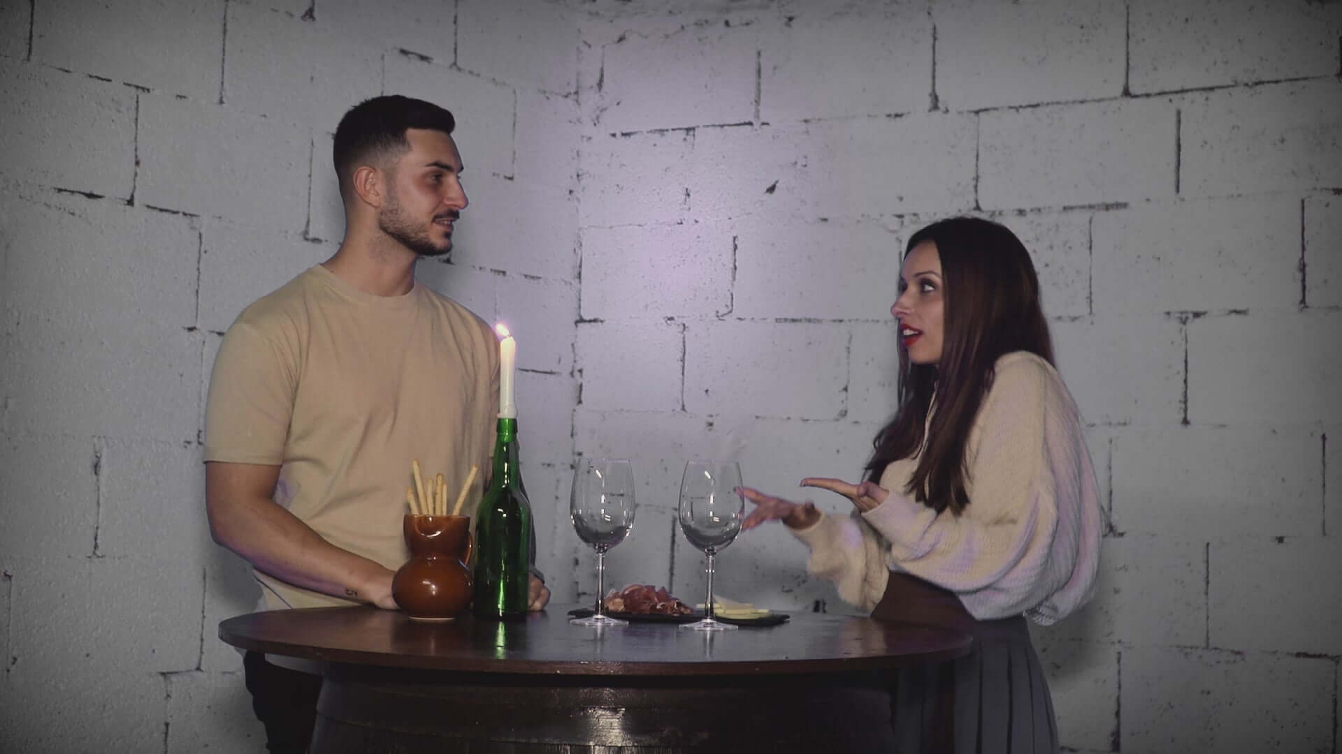 Cargar video: Pareja charlando en una mesa con botellas de vino y copas, en un ambiente contemporáneo y acogedor.